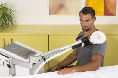 Seated patient receiving emFieldPro treatment over clothing at the shoulder, with the device and arm-mounted applicator visible.